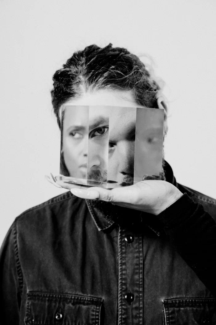 A person holds three angled mirror shards over their face, fragmenting their reflection into overlapping views.